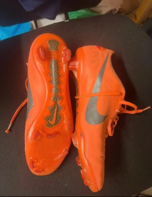 Nike orange fotbollsskor med dobbar - Säljer ett par grymma orange fotbollsskor från Nike med stora silvriga swoosh-loggor på sidorna. Skorna har snörning, låg profil och slitstark syntetisk ovandel. Dobbarna är utformade för bra grepp på planen och sulan har en cool detalj i kolfiber-look.