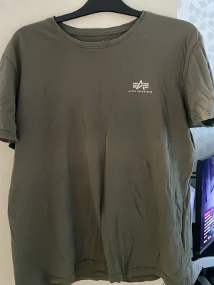 Olivgrön t-shirt Alpha Industries - Snygg olivgrön t-shirt från Alpha Industries med diskret logga tryckt på bröstet. Klassisk rund halsringning och korta ärmar. Perfekt basplagg till alla outfits och tillverkad i mjuk bomull för skön känsla.