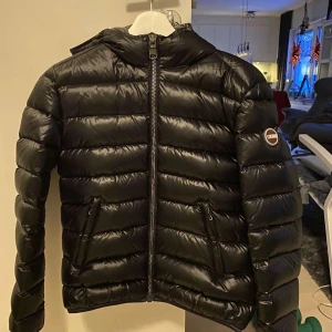 Svart pufferjacka från Colmar - Snygg svart pufferjacka från Colmar med huva och dragkedja framtill. Jackan har en quiltad design och är perfekt för kyligare väder. Den har en normal passform och långa ärmar. Storlek 8 USA vilket motsvarar ungefär 38.