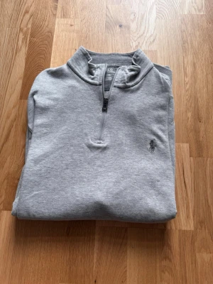 Grå halvzip från Polo Ralph Lauren - Snygg grå sweatshirt med halv dragkedja från Polo Ralph Lauren. Klassisk design med liten broderad logga på bröstet och hög krage. Perfekt till jeans eller chinos för en clean och avslappnad stil.