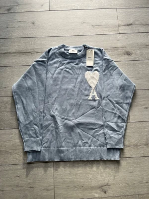 Ljusblå stickad tröja från Ami - Säljer en ljusblå stickad tröja från Ami med ett stort vitt hjärta och bokstaven 'A' broderat på bröstet. Tröjan har rund halsringning, ribbade muddar och avslut. Perfekt för dig som gillar stilrena detaljer och mjuka material.