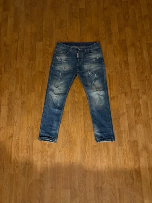Blå slitna dsquared2 jeans - Säljer ett par blå dsquared2 jeans med coola slitningar och distressed detaljer framtill. Jeansen har raka ben, klassisk femficksmodell och en tvättad look som ger en avslappnad vibe. Perfekta för dig som gillar streetwear och en trendig stil.