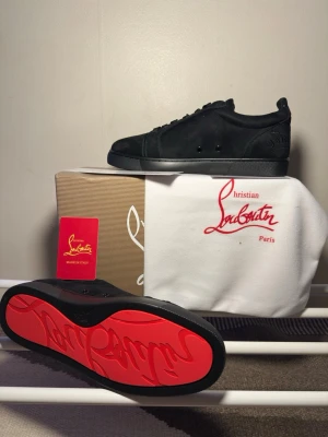 Christian Louboutin Skor - Snygga svarta sneakers från Christian Louboutin i mocka med ikonisk röd sula och diskret logga på hälen. Klassisk låg modell med svarta skosnören och stilren design. Perfekta för dig som vill sticka ut med exklusiva detaljer och streetstyle-vibe.