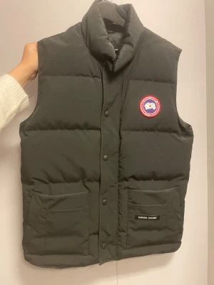 Svart dunväst från Canada Goose - Snygg svart dunväst från Canada Goose med klassisk logga på bröstet. Materialet är slitstarkt och fyllningen består av dun. Äkta väst, inga skam bud! Storlek S. Använd 2 gånger.