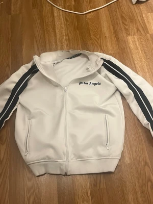Vit tracksuit från Palm angels  - Säljer en vit  tracksuit från Palm Angels med svarta ränder längs ärmarna och logga på bröstet. Jackan har dragkedja framtill, hög krage och två fickor med dragkedja. Perfekt för en sportig och trendig look. Säljer även byxorna  priset kan diskuteras så tveka inte att skriva!