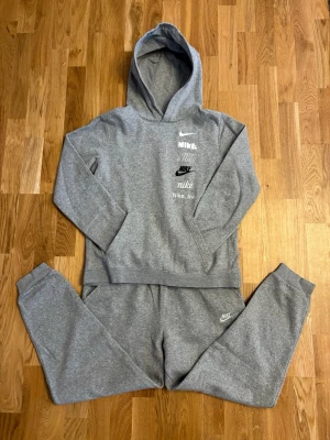 (BARN)Grå hoodie och byxor från Nike med tryck - Snygg grå hoodie och byxor från Nike med flera olika Nike-loggor. Hoodien har en klassisk huva och en stor magficka framtill. Tillverkad i mjukt sweatshirtmaterial som är perfekt för en avslappnad stil.