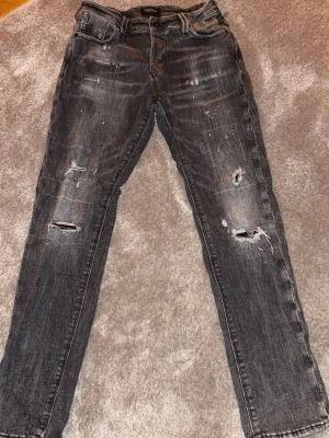 Grå slitna jeans med hål - Säljer ett par grå jeans med slitningar och hål på benen för en cool, avslappnad look. Jeansen har raka ben, klassiska fem fickor och detaljerade slitningar framtill och baktill. Perfekta för dig som gillar streetstyle och en edgy vibe. Passar mindre:)