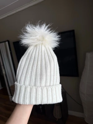 Vit ribbstickad mössa med pälsboll - Säljer en vit ribbstickad mössa med glitter i och en fluffig pälsboll på toppen. Oanvänd 