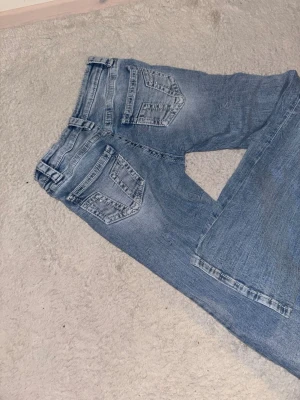 Ljusblå low waist bootcut jeans med detaljer på backfickorna - Säljer ett par ljusblå jeans som är low waist och bootcut ben och snygga sömdetaljer på bakfickorna. Jeansen har klassisk femficksdesign och är tillverkade i denim med lätt tvättad look. Perfekta för en avslappnad och trendig stil.