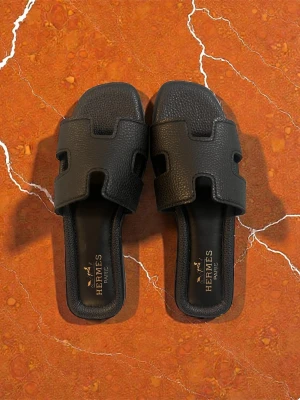 Svarta sandaler från Hmès - Svarta sandaler från Hmès med ikonisk H-design på ovandelen. Tillverkade i strukturerat läder med öppen tå och platt sula. Märkeslogga på innersulan och Eifel-tornet präglat under sulan. Perfekta för en trendig och avslappnad look.