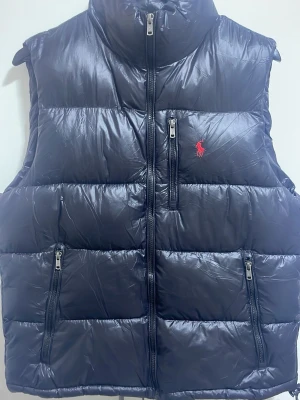 Svart dunväst från Ralph Lauren - Snygg svart dunväst från Ralph Lauren med hög krage och tre dragkedjeförsedda fickor framtill. Västen har det klassiska röda Polo-logot på bröstet och quiltad design som ger en puffig look. Perfekt för lager-på-lager och streetstyle. Aldrig använt!