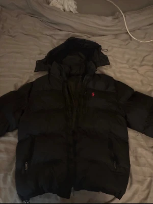 Svart pufferjacka från Polo Ralph Lauren - Säljer en svart pufferjacka från Polo Ralph Lauren med huva och dragkedja. Jackan har två sidofickor med dragkedja och den klassiska röda loggan på bröstet. Perfekt för kalla vinterdagar och riktigt skön att bära.