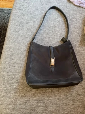 Svart axelremsväska med silverdetalj - Stilren svart handväska i mjukt material med axelrem och snygg silverfärgad spänndetalj framtill. Väskan har en klassisk, något triangulär form och minimalistisk design som passar till många olika looks.