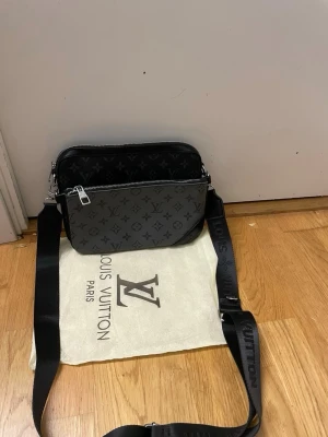 Louis Vuitton svart axelväska - Snygg svart axelväska från Louis Vuitton med klassiskt monogrammönster i grått och svart. Väskan har justerbar axelrem i textil med Louis Vuitton-logga, silverfärgade detaljer och dragkedja. Perfekt för dig som vill ha en stilren och lyxig accessoar.