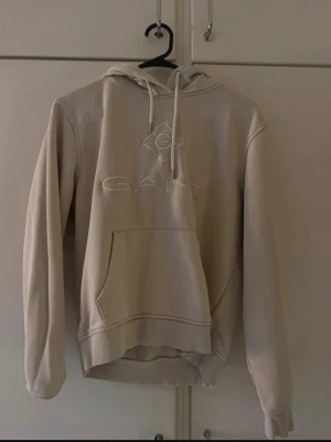 Beige hoodie från GANT med logga - Snygg beige hoodie från GANT med broderad logga på bröstet och klassisk känguruficka. Hoodien har dragsko i huvan och ribbade muddar vid ärmslut och nederkant. Perfekt för en chill och stilren look.