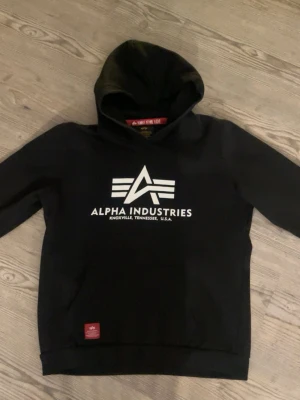 Svart hoodie från Alpha Industries - Superskön hoodie från Alpha Industries med stor vit logga och text på bröstet. Klassisk huva och känguruficka framtill. Använd fåtal gånger 💕