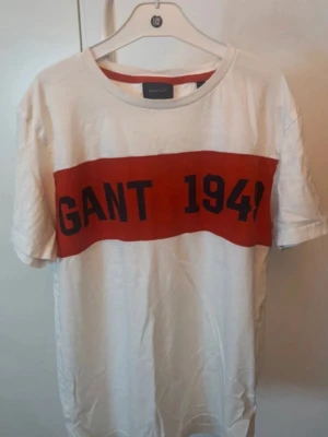Vit GANT t-shirt med röd logga - Vit t-shirt från GANT med en bred röd rand över bröstet och svart text 'GANT 1949'. 