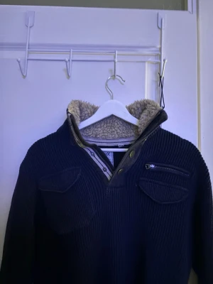 Mörkblå stickad tröja - Mörkblå stickad tröja med hög fodrad krage. Tröjan har dragkedja och knappar vid halsen, samt två bröstfickor varav en med dragkedja. Perfekt för kalla dagar och riktigt snygg till jeans. Skriv privat ifall du vill veta märket 