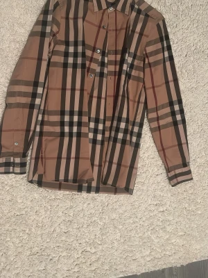 Skjorta från Burberry Brit ” sökes byten” - Säljer en beige rutig skjorta från Burberry Brit med klassiskt mönster i svart, vitt och rött. Skjortan har långa ärmar, knappar framtill och en snygg krage. Tillverkad i bomull för en skön känsla och stilren look. Äkta burberry använd 1 gång för ett bröllop. Låda finns, skriv om det är någon fråga eller fundering.