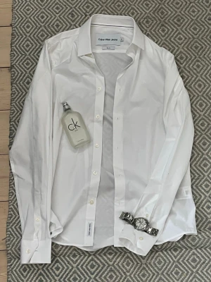 Vit skjorta Calvin Klein Jeans  - Klassisk vit skjorta från Calvin Klein Jeans (ALDRIG ANVÄNT) köpt för 1500. Skjortan har en stilren design som passar till allt. Tillverkad i mjuk bomull för en bekväm känsla och snygg passform. Grisch stil, perfekt för kalas eller speciella tillfällen. Kontakta om intresse! (OBS: accessoarerna i bild 1 ingår inte!)