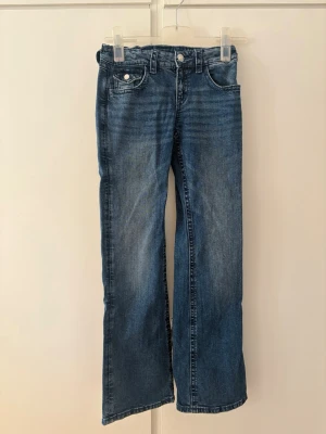 Blå bootcut jeans med broderade fickor - Säljer ett par blå bootcut jeans i klassisk denim med snygga slitningar framtill. Jeansen har fem fickor och bakfickorna är dekorerade med broderade rosetter och silverfärgade knappar. Perfekta för en avslappnad och trendig look. Samt är stretchiga 