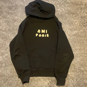 Svart hoodie från AMI Paris - Snygg svart hoodie från AMI Paris med broderad text i beige på bröstet. Klassisk passform med huva och ribbade muddar vid ärmslut och nederkant. Perfekt för en avslappnad och trendig look.