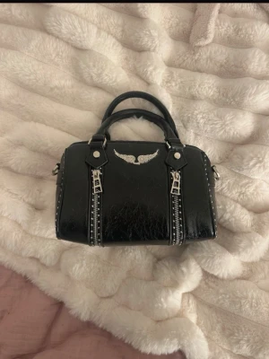 Svart handväska med silverdetaljer - Cool svart handväska i glansigt konstläder med två handtag, silverfärgade dragkedjor framtill och en liten vinge i metall som dekoration. Väskan har nitar längs kanterna och ett rymligt fack med innerfickor. Perfekt för dig som gillar en edgy stil.