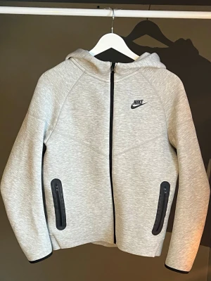 Ljusgrå Nike Tech med dragkedja - Snygg ljusgrå hoodie från Nike Tech med svart logga på bröstet. Hoodien har hel dragkedja framtill, två svarta dragkedjeförsedda fickor och en skön huva. Tillverkad i mjukt material som känns sportigt och modernt. Perfekt för en avslappnad streetwear-look. Det är i barnstorlek, så står 150 - 170, stämmer överens med mått på XS. Köpt från Nike, använd fåtal gånger 🩶