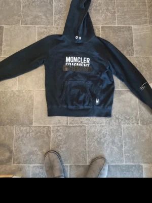 Svart Moncler Fragment hoodie - Snygg svart hoodie från Moncler Fragment med vit text på bröstet och klassisk känguruficka framtill. Huva med dragsko och ribbade muddar. Perfekt för en streetwear-look och enkel att matcha med jeans eller joggers.