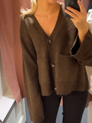 Brun kofta med fickor från Gina Tricot - Mysig brun kofta från Gina Tricot med v-ringning och stora knappar framtill. Koftan har två praktiska fickor och ribbade muddar vid ärmslut och nederkant. Perfekt för lager på lager och enkel att matcha med olika outfits.