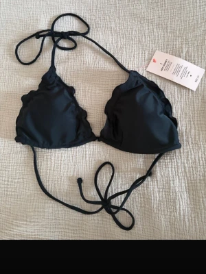 Svart bikiniöverdel med volangkant - Stilren svart bikiniöverdel från Nelly med volangkant längs kanterna. Klassisk trekantsmodell med knyt i nacke och rygg. Perfekt för sommaren och strandhäng!