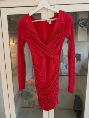 Röd omlottklänning med drapering - Säljer en röd kort omlottklänning från Gina Tricot med långa ärmar och snygg drapering framtill. Klänningen har en djup v-ringning och är tillverkad i ett mjukt, stretchigt material som sitter snyggt på kroppen. Perfekt för dig som vill sticka ut!
