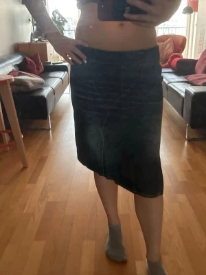 Svart långkjol i denim - Säljer en svart långkjol i denim med markerade sömmar och lätt asymmetrisk nederkant. Kjolen har en klassisk midja och enkel design som passar till många olika stilar. Perfekt för dig som gillar en stilren och tidlös look.