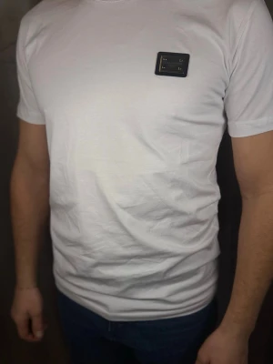 Vit Dolce & Gabbana t-shirt - Vit t-shirt från Dolce & Gabbana med korta ärmar och en snygg metallbricka med logga på bröstet. Klassisk passform och tillverkad i mjuk bomull som känns skön mot huden. Perfekt för dig som gillar stilrena plagg med exklusiva detaljer.
