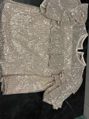 Beige paljettblus med volanger - Supersnygg beige blus täckt med glittrande paljetter och volangdetaljer framtill och på ärmarna. Blusen har rund halsringning och korta puffärmar som ger en trendig look. Perfekt för dig som vill sticka ut! Materialet känns lätt och skimrande.