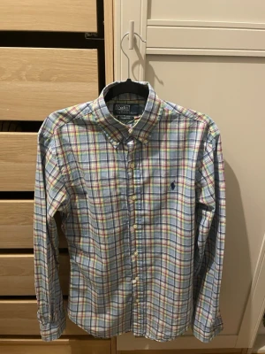 Rutig skjorta från Polo Ralph Lauren - Klassisk rutig skjorta från Polo Ralph Lauren med button-down krage och broderad logga på bröstet. Skjortan har långa ärmar och knäppning framtill. Färgerna är en mix av blå, grön, röd och gul på vit botten. Perfekt för en avslappnad men stilsäker look.