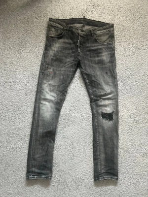 Grå slitna jeans med patch och färgstänk - Säljer ett par grå jeans med skinny fit och coola slitningar. Jeansen har en svart patch med nitar på ena benet och små färgstänk för en edgy look. Femficksmodell och dragkedjegylf. Perfekta för dig som gillar streetstyle och unika detaljer. har sytt för att det har gått sönder lite men det syns inte visas på bilden