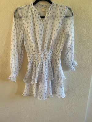 Vit blommig kortklänning med volang - Söt vit kortklänning från Gina tricot med blått blommönster, långärmad och med smock i midjan samt volangdetaljer på kjolen. Klänningen har v-ringning. Perfekt för sommarens alla tillfällen.
