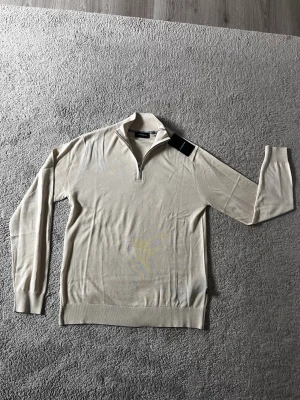 Beige halvzip-tröja från Lindbergh - Snygg beige tröja från Lindbergh med halvzip och ståkrage. Tillverkad i mjukt, tunt material som passar perfekt för lager-på-lager. Långa ärmar och ribbade muddar vid ärmslut och nederkant ger en clean look.