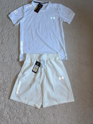 Vitt träningsset från Under Armour - Snyggt vitt träningsset från Under Armour med t-shirt och shorts. Båda plaggen har logga och textdetaljer i vitt. T-shirten har rund hals och korta ärmar, shortsen har resår i midjan och är knälånga. Perfekt för gym eller löpning.