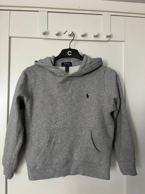 Grå hoodie från Polo Ralph Lauren - Klassisk grå hoodie från Polo Ralph Lauren med huva och känguruficka. Liten mörkblå logga broderad på bröstet. Tillverkad i mjukt sweatshirtmaterial, perfekt för en avslappnad stil.