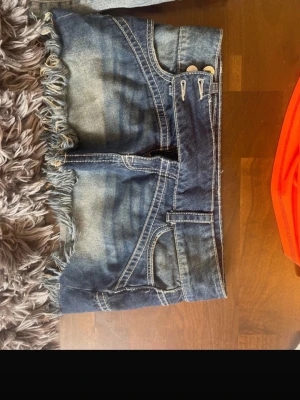 Blå jeanskjol med fransar - Säljer en blå jeanskjol i kort modell med råa fransar längs nederkanten. Kjolen har klassiska fickor fram och bak, samt knapp- och dragkedjestängning. Perfekt för dig som gillar en avslappnad och trendig stil.