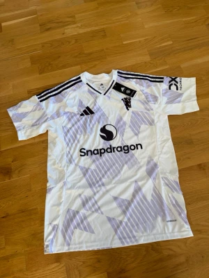 Vit Adidas fotbollströja med mönster - Snygg vit fotbollströja från Adidas med ljuslila geometriskt mönster och svarta detaljer. Tröjan har korta ärmar, svarta ränder på axlarna och Snapdragon-logga på bröstet. Tillverkad i lätt och ventilerande material, perfekt för träning eller match.