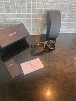 Svarta solglasögon från Prada - Snygga svarta solglasögon från Prada med klassisk rektangulär form och diskreta röda detaljer på skalmarna. Kommer med originalfodral, box och äkthetskort. Perfekta för dig som vill ha en stilren och exklusiv accessoar.