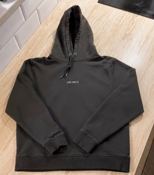 Mörkgrå hoodie från Les Deux - Snygg mörkgrå hoodie från Les Deux med broderad logga på bröstet. Klassisk modell med huva och dragsnören samt känguruficka framtill. Tillverkad i mjukt bomullsmaterial och har ribbade muddar vid ärmslut och nederkant. Perfekt för en avslappnad stil.