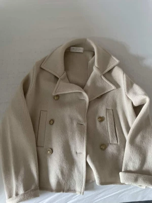 Beige ullkappa från Busnel - Stilren beige ullkappa från Busnel med dubbelknäppning och guldiga knappar. Klassisk krage och raka fickor framtill. Perfekt för dig som gillar tidlös design och vill ha en snygg och enkel kappa till garderoben. helt ny