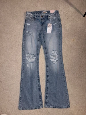 Ljusblå bootcut jeans med slitningar - Säljer ett par ljusblå bootcut jeans med tydliga slitningar framtill och på bakfickorna. Jeansen har klassisk femficksmodell, hällor för skärp och en avslappnad passform. Materialet är denim i bomull och de har en trendig vintagekänsla.
