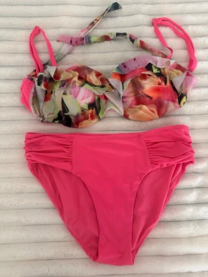 Färgglad bikini med blommigt mönster - Säljer en snygg bikini med blommigt mönstrad överdel i flera färger och enfärgad rosa underdel. Överdelen har vadderade kupor och knyt i nacken. Materialet känns mjukt och stretchigt, perfekt för lata dagar på stranden.