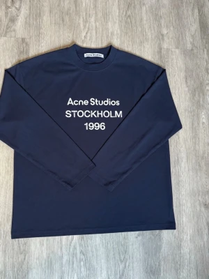 Acne studios long sleeve - Snygg mörkblå långärmad tröja från Acne Studios med vit text 'Acne Studios STOCKHOLM 1996' på bröstet. Klassisk rund halsringning och rak passform. Perfekt för dig som gillar stilrena och trendiga plagg.