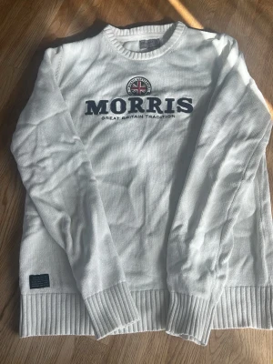 Vit stickad tröja från Morris - Vit stickad tröja från Morris med rund halsringning och ribbade muddar. Framtill finns ett broderat Morris-emblem med Union Jack och texten 'Great Britain Tradition' i marinblått. Perfekt för dig som gillar klassisk stil.